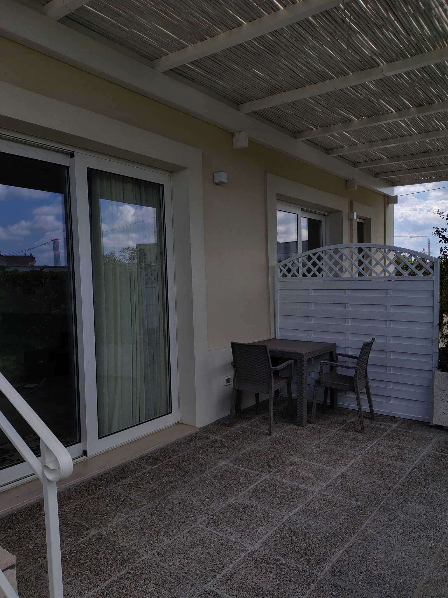 A covered patio area with a table and chairs, a sliding glass door, and a white fence.