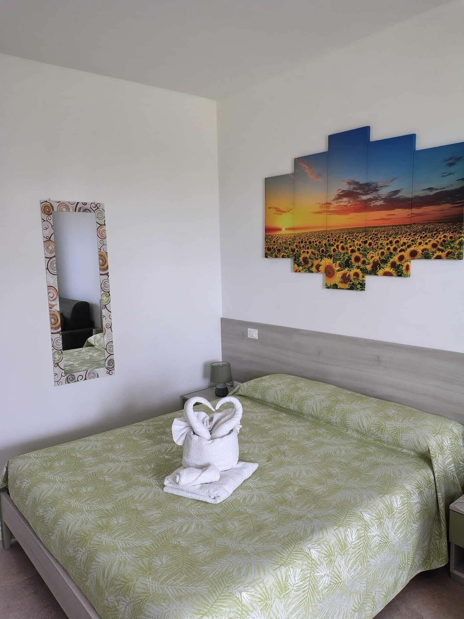Een opgemaakt bed met een groen bedovertrek, een witte handdoek met een hartvorm, een spiegel met decoratieve lijst en een zonsondergangschilderij aan de muur.