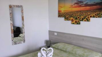 Een opgemaakt bed met een groen bedovertrek, een witte handdoek met een hartvorm, een spiegel met decoratieve lijst en een zonsondergangschilderij aan de muur.