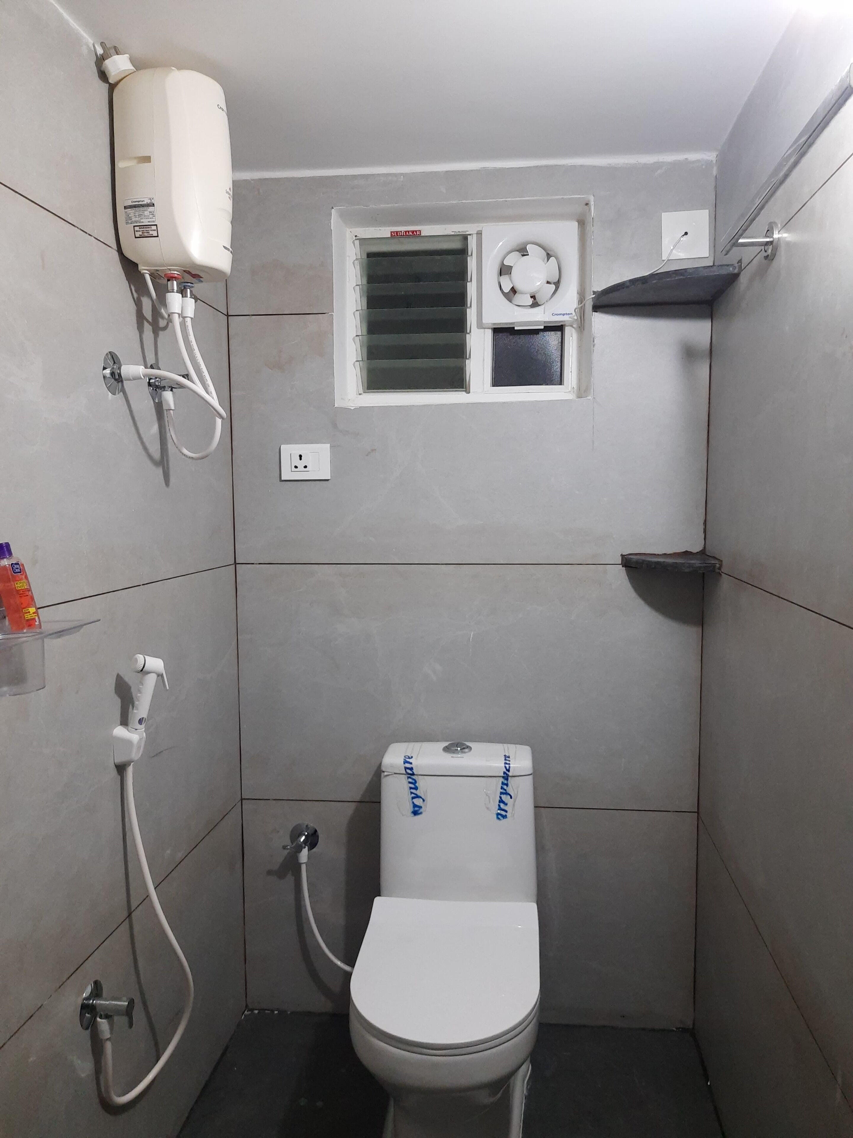 Une petite salle de bain carrelée, équipée de toilettes, d’une fenêtre avec stores, d’un radiateur mural et d’un pommeau de douche.