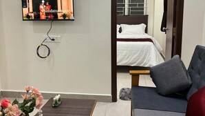 A hotel room with a bed, a sofa, a TV mounted on the wall, a coffee table with a vase of flowers, and a door leading to another room. - PS ELITE HOME STAY (Tirupati)