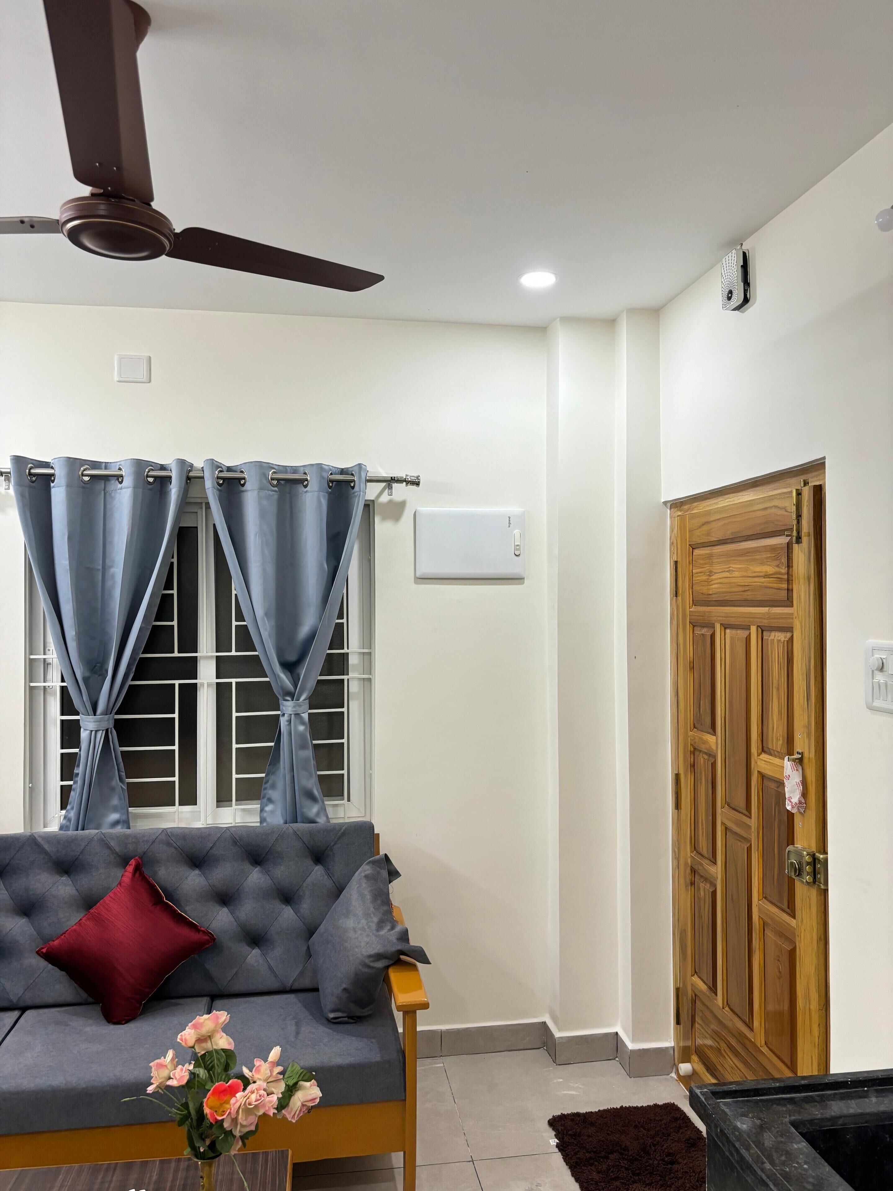 Une pièce avec un canapé gris, un ventilateur de plafond, une fenêtre avec des rideaux, une porte en bois et un thermostat fixé au mur.