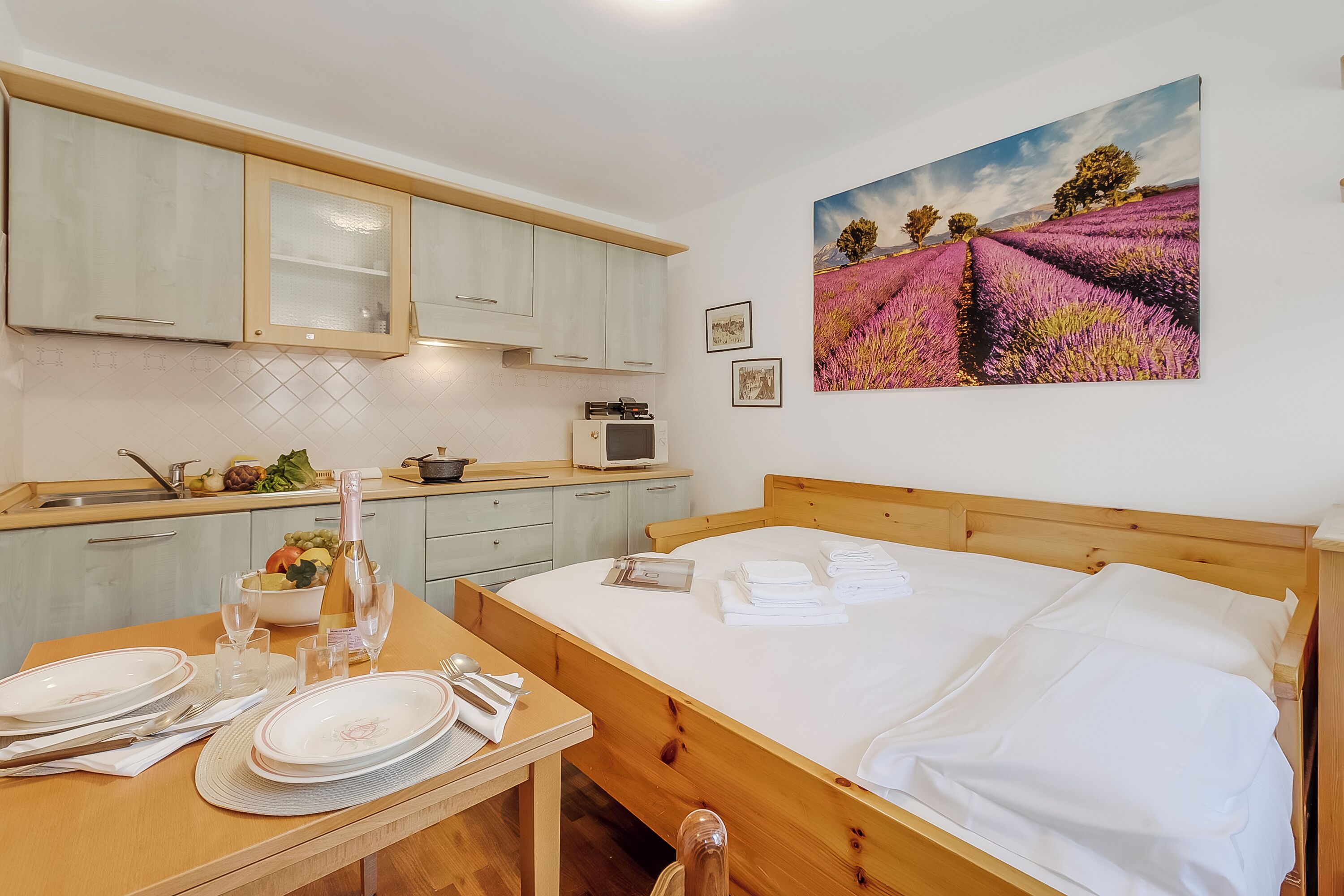 Une chambre avec un lit en bois, une table à manger dressée avec des assiettes et des couverts, une cuisine avec des meubles de couleur claire, et un tableau représentant un champ de lavande accroché au mur.