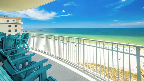 A balcony with teal Adirondack chairs overlooking a beach with a white railing and a pier in the distance.
