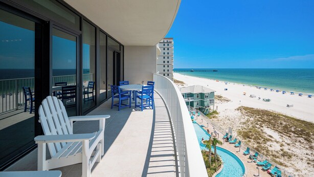 A balcony with a view of the beach, a pool, and a high-rise building in the background.