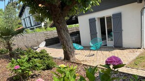 A garden with a tree, a patio area with chairs, and a house with a white exterior and grey shutters.