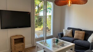 A modern living room with a flat-screen TV mounted on the wall, a glass-top coffee table, a dark sofa with patterned cushions, and a wicker pendant light.