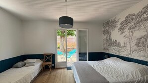 A bedroom with a large bed, a smaller bed, a chair, a table, and a view of the outdoors through a sliding door.