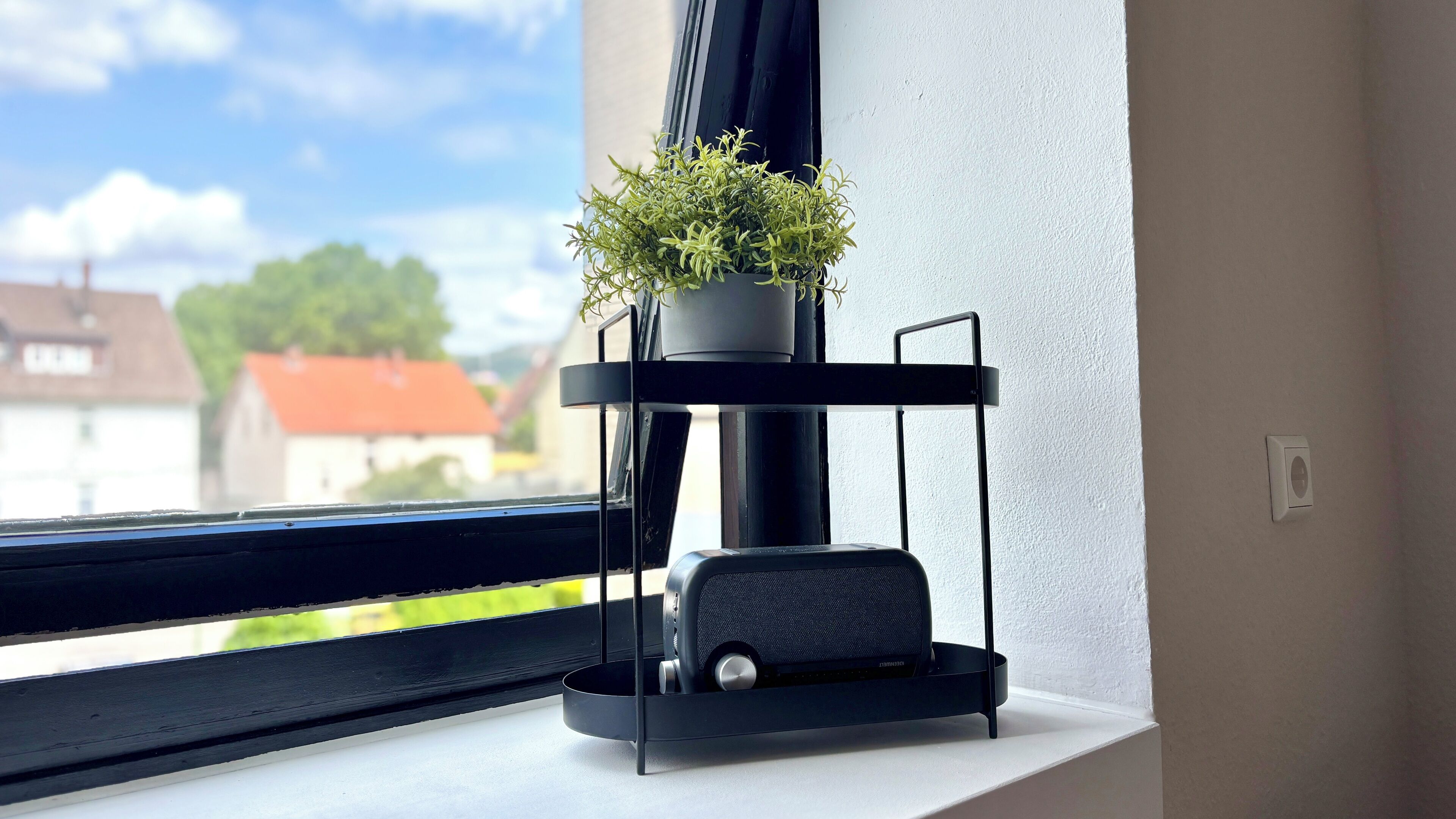 Un haut-parleur noir est posé sur une étagère blanche, près d’une fenêtre, et une plante en pot se trouve à proximité.