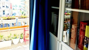 A balcony with potted plants, a view of buildings, and a bookshelf with books.