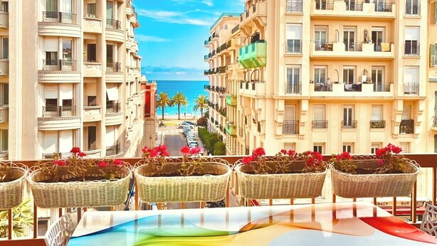 A balcony with wicker chairs and a table, overlooking a coastal street with multi-story buildings and a clear blue sky.