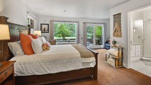 A bedroom with a large bed, a wooden nightstand, a desk, a chair, a window with a view of greenery, and a sliding glass door leading to an outdoor area. - Yaya’s: 4BR Retreat | Lake • Golf • Ski (Tahoe Vista)
