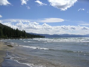 Ein Strand mit Wellen, einem Steg und einer bewaldeten Hügelkette im Hintergrund.