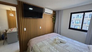 Ein Hotelzimmer mit Bett, Wandfernseher, Klimaanlage, Fenster mit Blick auf Gebäude und einem kleinen Schreibtisch mit Stuhl.