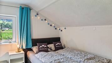 A bedroom with a bed, a nightstand, a lamp, a window with curtains, and a view of greenery outside.