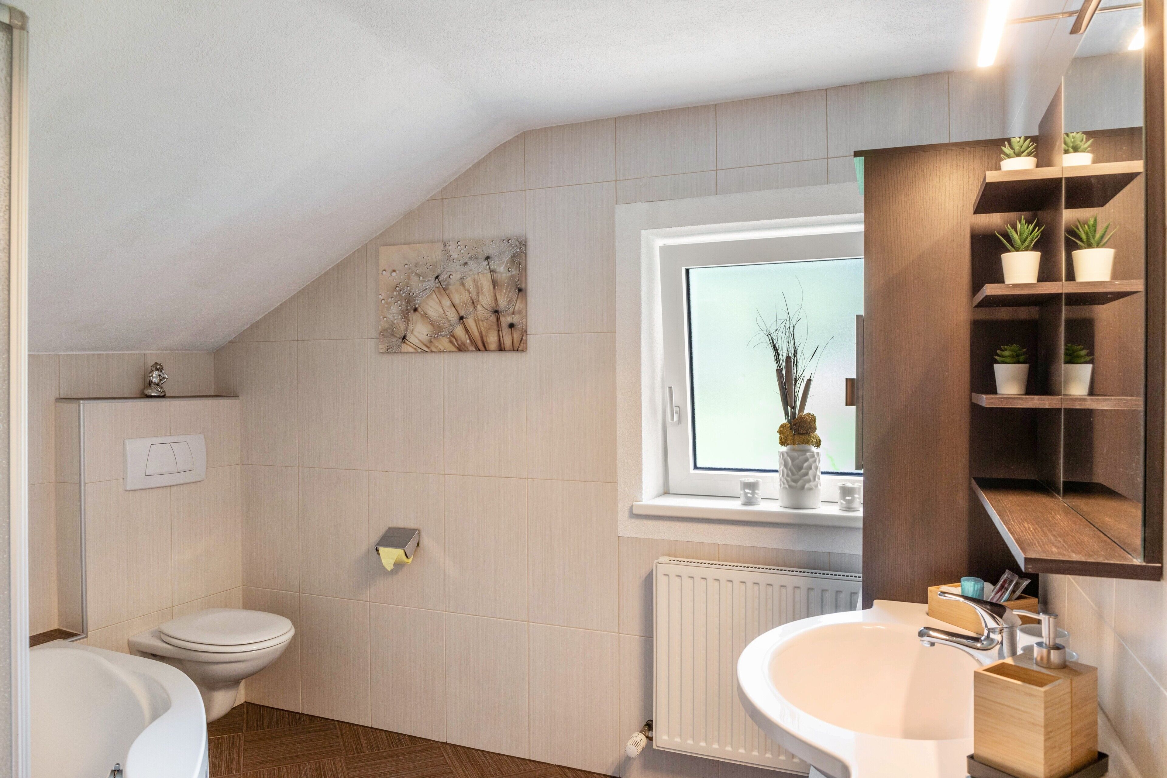 A modern bathroom with a toilet, bathtub, and sink, wooden floor, and a window with plants.