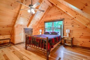 Een slaapkamer met houten lambrisering op zolder, met een bed, nachtkastje, plafondventilator en raam.