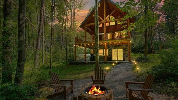 Une cabane dotée d’une terrasse, entourée de forêt, et un foyer avec des chaises.