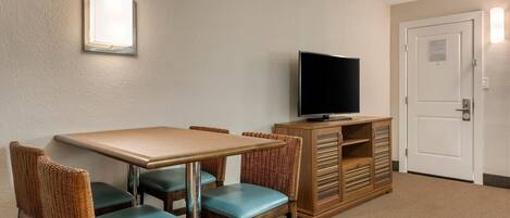 A hotel room with a dining table, four chairs, a television on a wooden cabinet, and a white door.