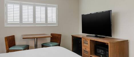 A hotel room with a bed, a TV, a desk, two chairs, and a window with blinds.