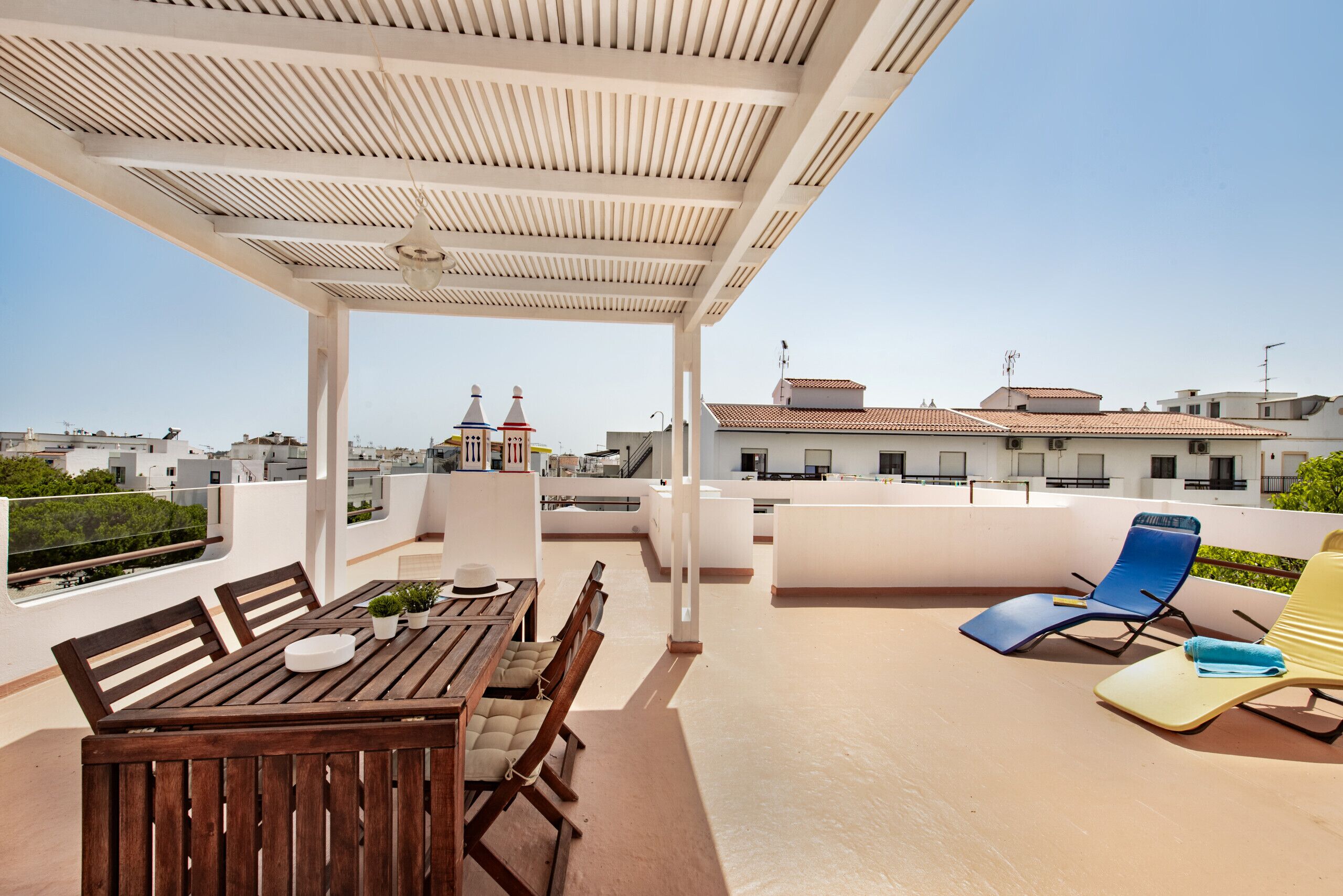 A rooftop terrace with a wooden table and chairs, a blue lounge chair, and a yellow lounge chair.