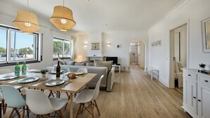 A spacious living area with a dining table set for four, a sofa, and a TV. There are large windows and a view of the outside. - Luzia Apartment in Tavira (Tavira)