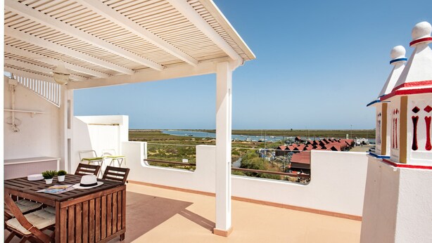 A covered outdoor area with a wooden table and chairs, a white railing, and a view of the sea and buildings in the distance. - Luzia Apartment in Tavira (Tavira)