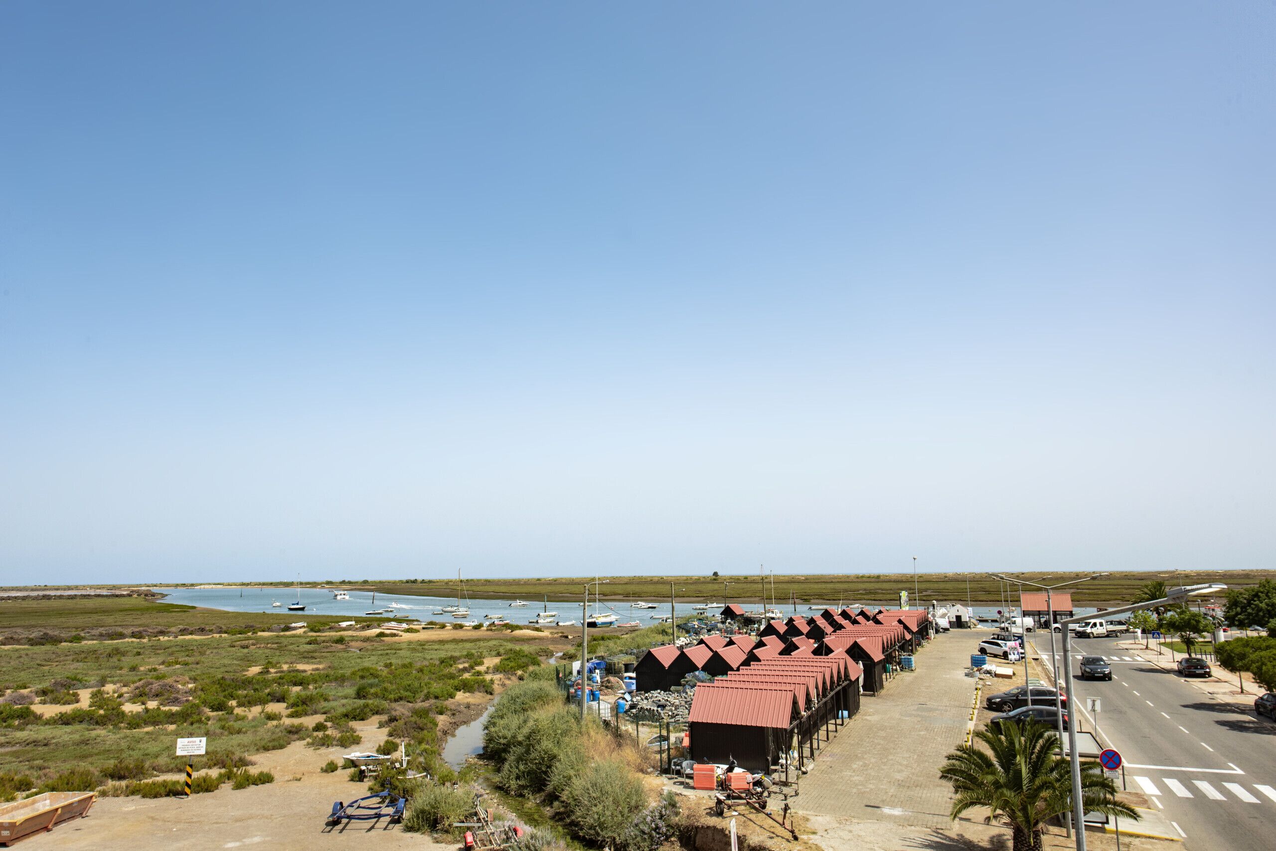 A coastal area with a row of red-roofed buildings, a road with vehicles, and boats on the water.