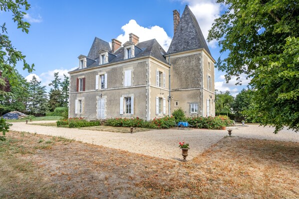 A large, historic building with multiple stories, a prominent tower, and a well-maintained garden with pathways and flower beds.