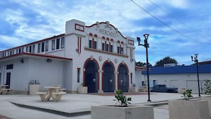 Un bâtiment à deux étages, doté d’une façade blanche et de bordures rouges, avec une grande entrée en arc de cercle et une enseigne sur laquelle on peut lire « Mercado ».