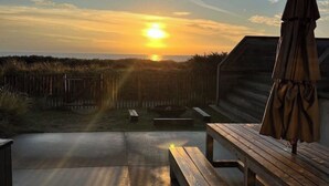 Vista del atardecer desde una terraza de madera con un banco y una sombrilla, con vista a una playa y una valla de madera.