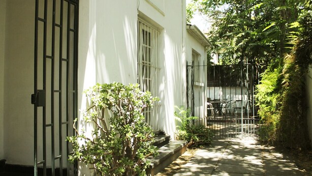 A narrow pathway leading to a white building with barred windows and a small garden area with a potted plant. - Casa y patio en mejor barrio de Santiago (Santiago)