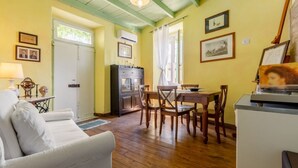A room with a wooden floor, a white sofa, a dining table with chairs, a kitchen area with a stove, and framed pictures on the walls.