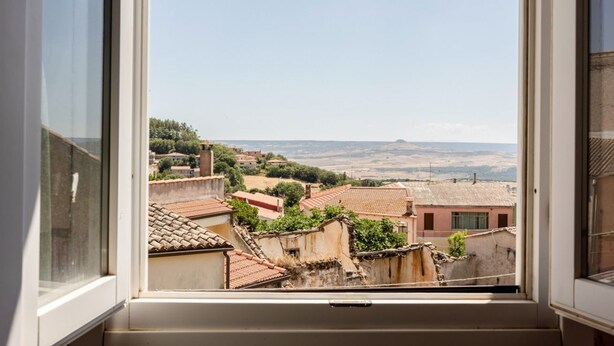 A window view of a residential area with terracotta-roofed buildings, a clear sky, and distant hills.