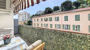 Un balcon avec un auvent à rayures, une table recouverte d’une nappe à motifs et un pot de fleurs.