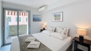 Une chambre d’hôtel avec un lit, deux oreillers, une table de chevet avec une lampe, un tableau au mur et un balcon donnant sur des immeubles.