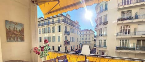 A balcony with a yellow awning, wicker furniture, a table with a vase of flowers, and a painting on the wall.