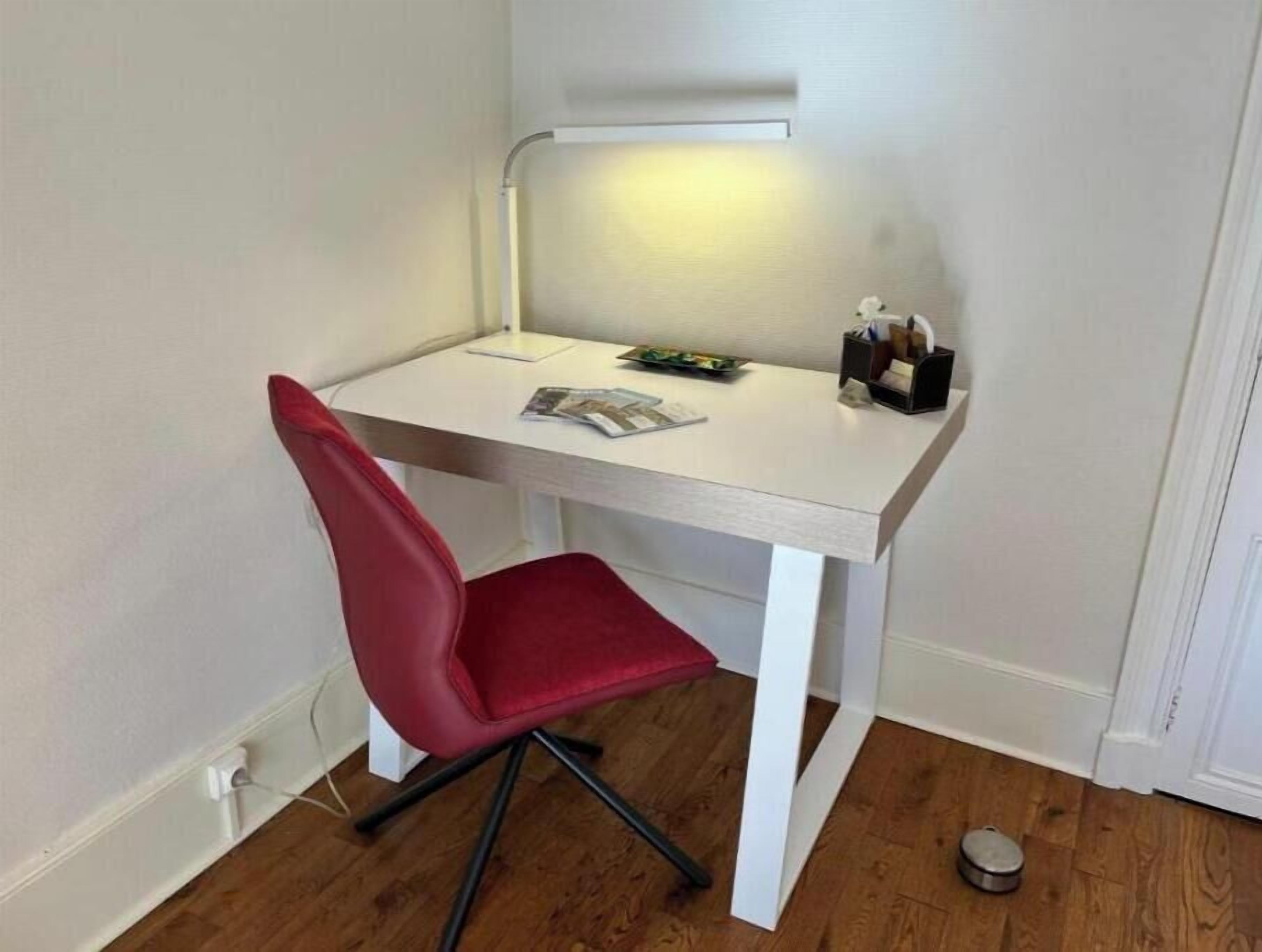 A small, white desk with a red chair, a lamp, and some items on the desk.