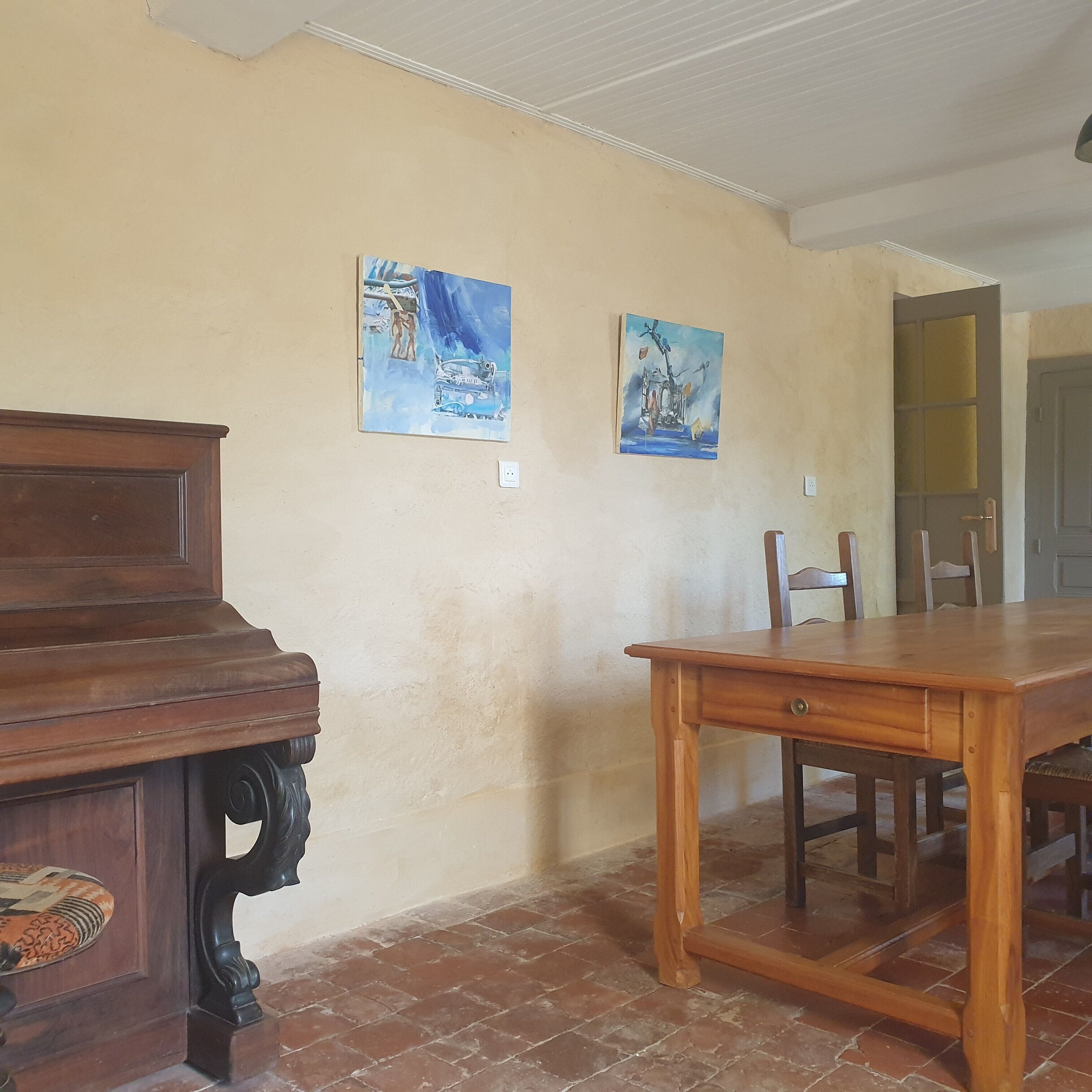 Una habitación con un piano de madera, una mesa de comedor y dos cuadros colgados en la pared.