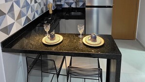 Una cocina moderna con encimera negra, un refrigerador de acero inoxidable y un horno empotrado. Hay tres lámparas colgantes sobre la encimera y una silla negra con asiento de fibras trenzadas.