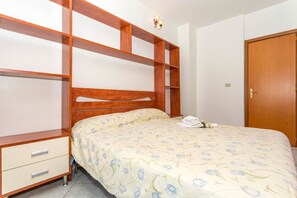 Une chambre à coucher avec un lit en bois, une table de chevet avec des tiroirs, une étagère fixée au mur et une porte.