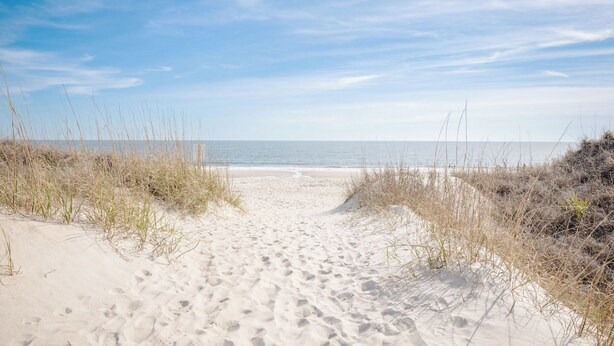 Un sentier de sable qui longe la plage et mène à l’océan, bordé de gazon et de dunes.