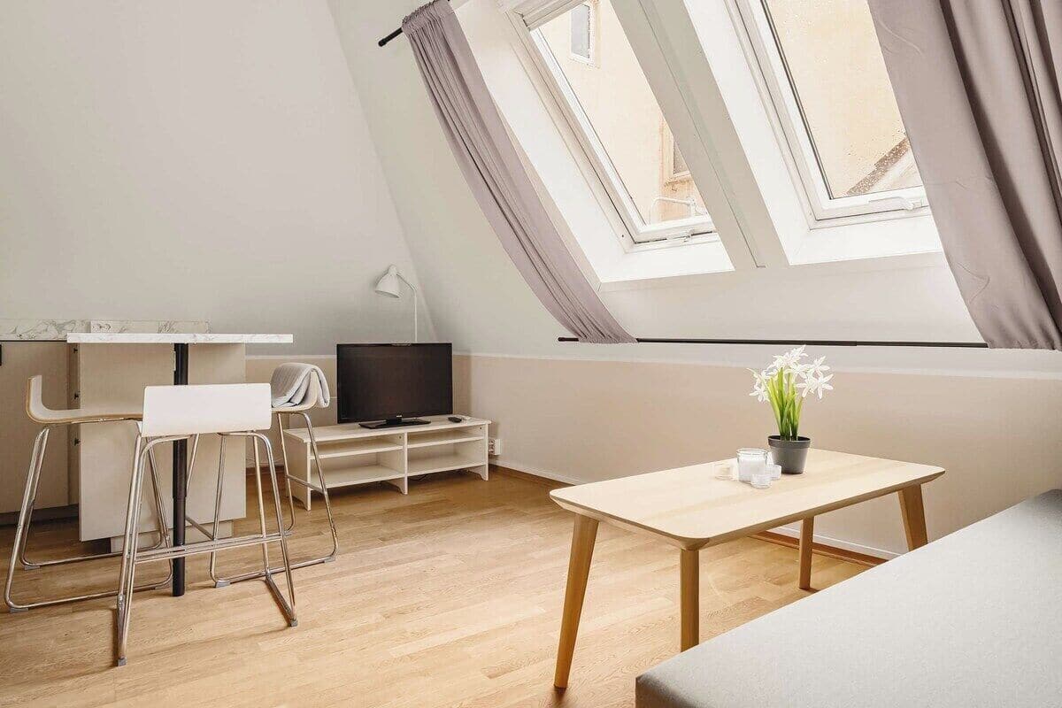 Une pièce moderne et bien éclairée, avec un plafond de style industriel, une petite table sur laquelle est posée une plante en pot, un téléviseur sur une étagère basse et deux tabourets de bar.