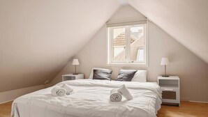 Una camera da letto con soffitto spiovente, un letto con biancheria bianca, due comodini con lampade e una finestra vista edifici.