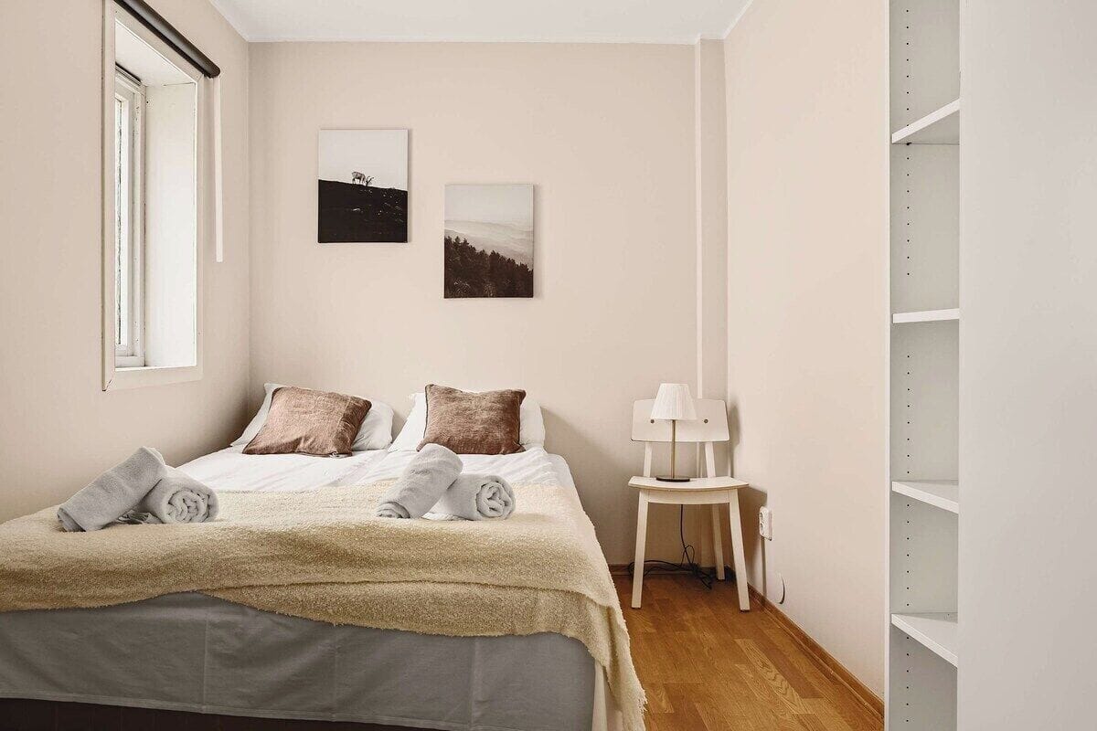 Un lit bien fait, recouvert d’une couverture beige et agrémenté de deux oreillers, une petite table de chevet blanche avec une lampe, et deux tableaux encadrés accrochés au mur.
