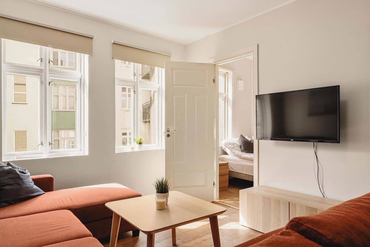 Un salon moderne avec un canapé, une petite table sur laquelle est posée une plante, un lit à l’arrière-plan et un téléviseur fixé au mur.