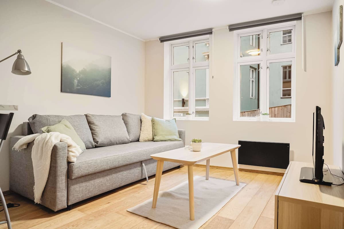 Un salon moderne avec un canapé gris, une petite table blanche et un téléviseur posé sur un meuble en bois.
