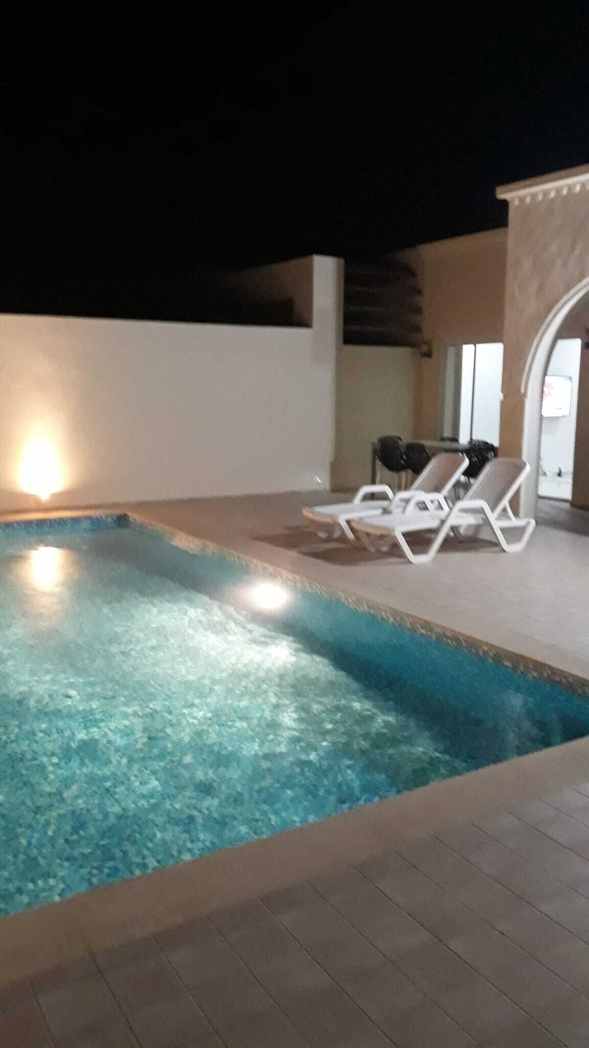 Una piscina interior con agua cristalina, rodeada de sillas reclinables blancas y un suelo de baldosas.