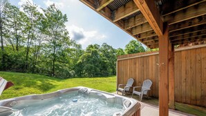 Ein Whirlpool unter einer Holzterrasse mit zwei Adirondack-Stühlen, umgeben von einem Rasen und Bäumen.