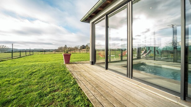 A modern outdoor pool area with a wooden deck, large glass windows, and a view of a green field. - The Vagastines (Saint-Germain-du-Plain)